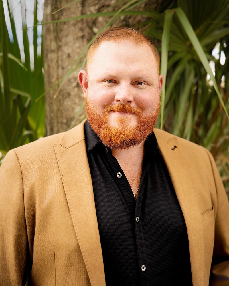 James Weber, a smiling man with red hair and a red beard, wearing a tan blazer over a black shirt, standing in front of palm trees.